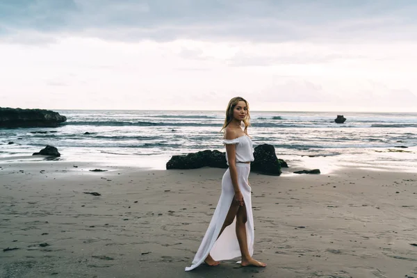 Beyaz elbiseli genç etnik kadın gezginin tüm vücudu dalgalı deniz kenarındaki kumlu sahil boyunca yürüyüp kameraya bakarken.