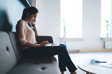 Klas kıyafetli genç etnik bayan yöneticinin koltukta otururken telefon konuşması ve dizüstü bilgisayarını karıştırmasının yan görüntüsü.