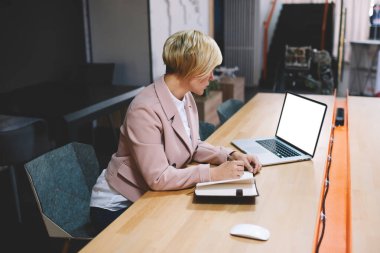Odaklanmış sarışın iş kadınının dizüstü bilgisayara göz atması ve modern ofisteki masada oturup not alması.