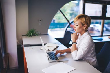 Modern ofiste masada oturup dizüstü bilgisayarla yazı yazarken beyaz gömlekli, çenesine dokunan kadın sekreterin yan görüntüsü.