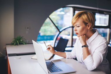 Ciddi kadın işçilerin resmi kıyafetleri içinde, kısa saçlı ve elleri çene kısmında masada oturmuş dizüstü bilgisayarla sörf yaparken yan görünümü.