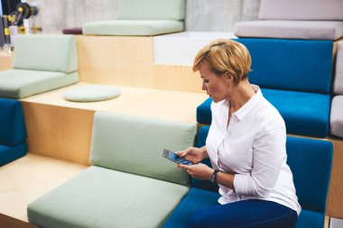 Beyaz tişörtlü, orta yaşlı, kısa saçlı, akıllı telefonlu, dinlenme odasında dinlenen bir kadının yan görüntüsü.