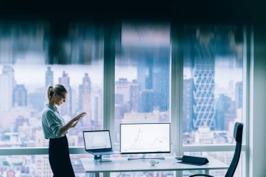 Gözlüklü tablet kadın ve resmi giyim senkronizasyon cihazlarının şehir merkezindeki modern iş yerindeki tam duvar penceresinin yanındaki ekrana odaklanmış görüntüsü