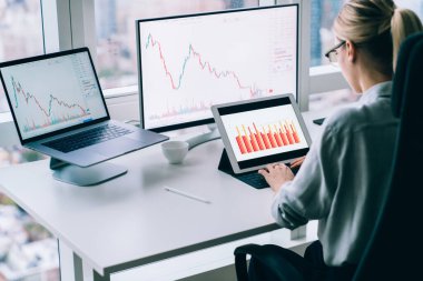 Resmi olarak tanınmayan bir kadının yüksek açılı arka görüntüsü modern ofiste masada oturuyor ve kripto para birimi bitcoin grafiği ile çalışıyor.