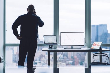 Pencere kenarında el pencerede dururken cep telefonuyla konuşan tanınmayan bir erkek yöneticinin arka planı ve ofiste çizelgeleri olan bilgisayarlar.