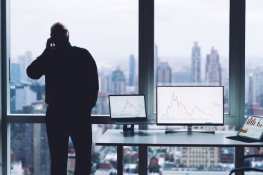 Cep telefonuyla konuşan tanınmayan bir erkek yönetici modern ofisteki çizelgelerle birlikte multimedya cihazlarının yanında el ele dururken...