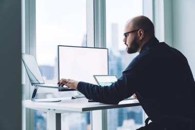 Ofisteki masadaki verileri analiz ederken tablet ve bilgisayarın yanında netbook izleyen genç sakallı finansörün yan görünüşü