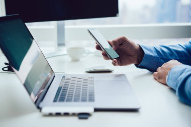Resmi giysiler içinde, cep telefonuna göz atan ve güncel çalışma alanında dizüstü bilgisayarla birlikte oturan isimsiz bir erkek yönetici.