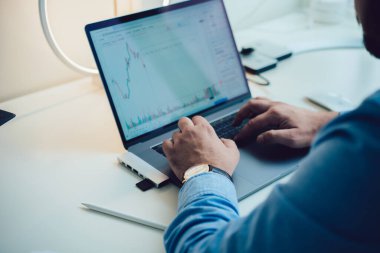 Resmi olarak tanınmayan erkek girişimcinin modern ofiste istatistiksel rapor oluştururken dizüstü bilgisayarda yazması yüksek açılı.