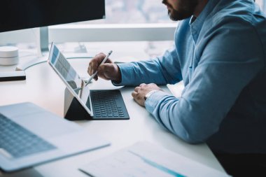 Modern ofiste bilgisayar ve dizüstü bilgisayarla masada otururken stilini kullanarak klavyeye yerleştirilmiş tablet üzerinde çalışan isimsiz sakallı erkek girişimcinin yan görüntüsü