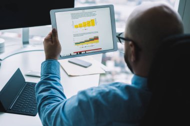 Tuş takımı ile masada oturan ve istatistiksel grafiklerle tablet ekranına bakan isimsiz erkek simsarın yüksek açılı arka görünümü