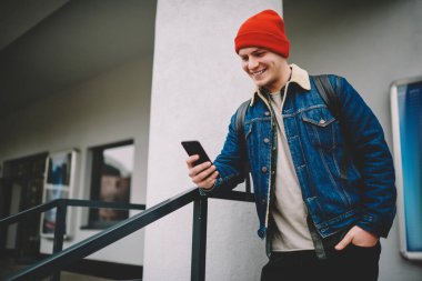 Cebinde cebi olan neşeli, yakışıklı bir erkeğin bina kenarında dururken arkadaşlarıyla sohbet etmek için cep telefonunu kullanarak korkuluklara dayanması.