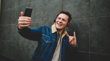 Geniş açılı, günlük kıyafetleri olan, kulaklıklı, duvarın yanında duran, akıllı telefon kullanarak selfie çeken, rock n roll hareketlerini gösteren genç bir erkek.