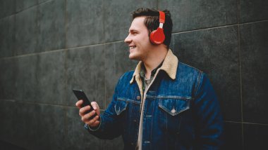 Geniş açılı, gündelik giysiler içinde, kablosuz kulaklıklı, boş zamanlarında cep telefonuyla duvarın yanında duran genç bir erkek.