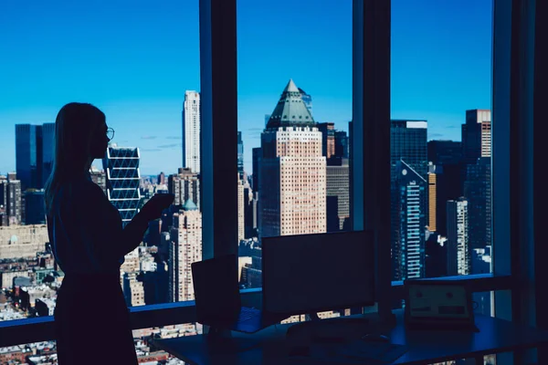 コンピュータとテーブルの近くに立つ女性労働者のシルエットの側面図とオフィスの窓から現代都市の景色を楽しむ — ストック写真