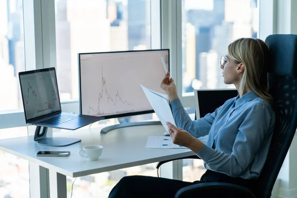 Bilgisayarda ve dizüstü bilgisayarda çalışırken modern ışık çalışma alanındaki kağıtlarla masa başında çalışan konsantre kadın girişimci.