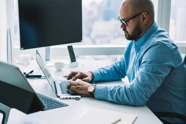 Yan görünüş erkek ofis çalışanı resmi gömlekli modern aletlerle masada oturuyor ve hafif iş yerinde dizüstü bilgisayarda klavye kullanıyor.