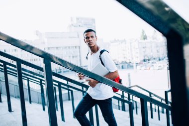 Kırmızı sırt çantalı genç bir erkek öğrencinin modern dizüstü bilgisayarı ve içecek paketini üst katta kentsel arka planda metal korkulukların yanında yürürken görüntüsü