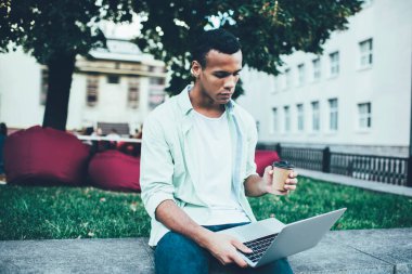 İnternette internette sörf yapan ciddi bir Afrikalı genç adam şehir parkında dinleniyor ve tek kullanımlık bardaktan sıcak içecek içiyor.