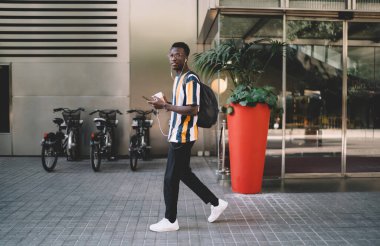 Milenyum turistinin tam boy portresi kahve ile şehir merkezinde yürümek ve kulaklıkla mobil müzik dinlerken kameraya bakmak, cep telefonlu Afrikalı Amerikalı hippi adam.