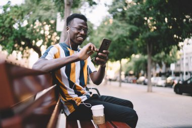 Görme düzeltmesi için gözlüklü mutlu hipster adam gülerek komik video bloğu izlerken cep telefonunu kullanıyor, kulaklıklı neşeli erkek blogcu sesli mesaj alıyor.
