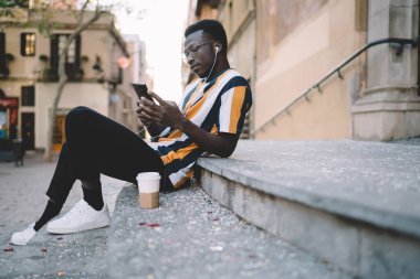 Milenyum hipster 'ının şehirde boş zamanlarında online rezervasyon ve bankacılık yapmak için akıllı telefon kullanmasının yan görüntüsü, kulaklık takan koyu tenli erkek blogcu mobil uygulama üzerinden çevrimiçi video bloğu izliyor.