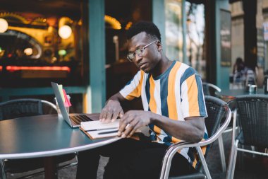 Modern dizüstü bilgisayar, serbest çalışma yoluyla proje araştırmaları için zaman harcarken eğitim ders kitabındaki bilgilendirme notlarını kontrol eden gözler için optik göz giysisi alanındaki yetenekli siyah öğrenci