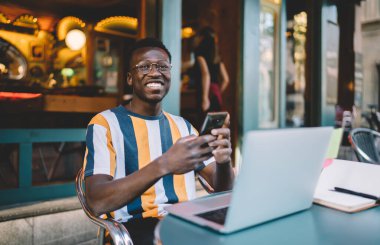 Dijital akıllı telefonlu neşeli erkek blogcu sokak kafesinde oturuyor ve kameraya gülümsüyor. Optik gözlüklü, koyu tenli hippi çocuk cep telefonu ve dizüstü bilgisayar kullanıyor.