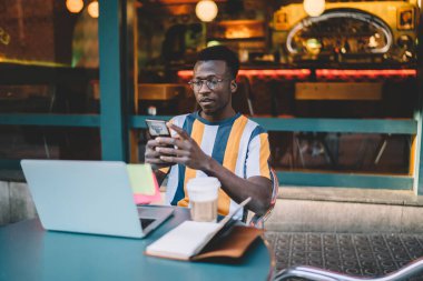 Millennial IT uzmanı gözlük kontrolünde, kaldırım caddesi kafede uzaktan çalışan modern cep telefonu cihazıyla ilgili tebligat aldı, Afrikalı Amerikalı blog yazarı içerik yayını okuyor