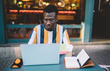 Program kodu üzerinde uzaktan çalışan Afrikalı Amerikalı erkek yazılım geliştirici web bilgilerini araştırmak için laptop teknolojisi kullanıyor. Netbook üzerinden 4G internete bağlanan gözlüklü siyahi adam.