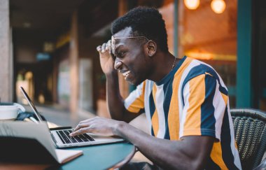 Şok olmuş, çizgili tişörtlü, alnına gözlük takan genç bir adamın yan görünüşü. Masada oturup laptopun inanılmaz sonuçlarını kontrol ederken.