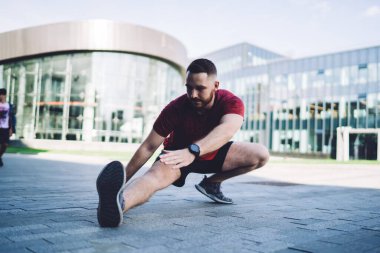 Aktif kıyafetli sporcu erkek, bulutsuz havada modern cam bina yakınlarında antrenmandan önce ısınırken uzanan bacaklara bakıyor.