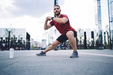 Spor kıyafetleri ve spor ayakkabıları giymiş, plastik su şişesinin yanında duran, spor yapmadan önce esneyen bacaklı, yoğun sakallı sporcu.