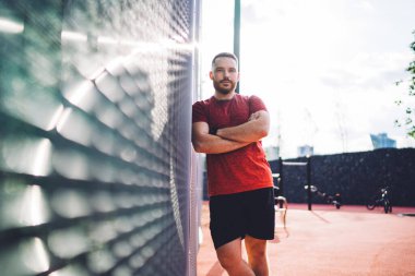 Spor kıyafeti giyen ciddi sakallı erkek sporcu kollarını kavuşturup çitin karşısında duruyor ve stadyumda antrenman yaptıktan sonra mola veriyor.