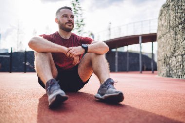 Ciddi sakallı, aktif giysili bir erkek yazın güneşli bir günde antrenmandan önce stadyumda oturuyor.