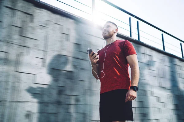 Smiling Male Red Shirt Shorts Looking Away Using Smartphone Earphones — стоковое фото