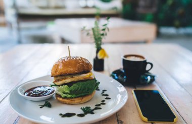 Ahşap masaya soslu lezzetli hamburger, bir fincan kahve ve gündüz kahveli siyah ekran akıllı telefon.