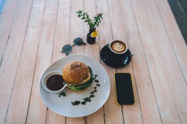Akıllı telefonun yukarısından, siyah ekranlı, lezzetli hamburgerli ve kafede bir fincan aromalı kahveyle.