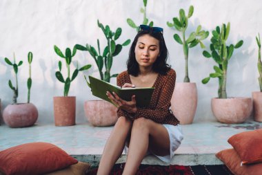 Güneşli avluda taş zeminde dinlenen ve deftere notlar yazan modaya uygun yaz kıyafetleri giyen güzel odaklanmış genç bir bayan.
