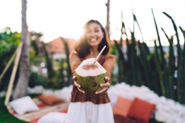 Yazlık kıyafetleri içinde, mutlu bir şekilde gülümseyen ve egzotik otellerin terasında samanla hindistan cevizi suyu içen bulanık genç bayan.