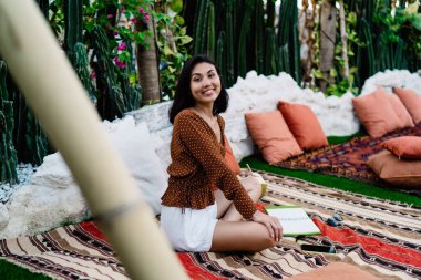 Sıska, etnik bir kadın battaniyeye sarılıp kameraya dostça gülümseyerek bakarken bacak bacak bacak üstüne atmış bir şekilde defterin önünde oturuyor.