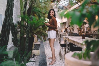 Alışveriş merkezinde yeşilliğin yanında dikilirken bilgi ve mesajlaşma için akıllı telefonu kullanan gülümseyen, narin, etnik bir kadın.