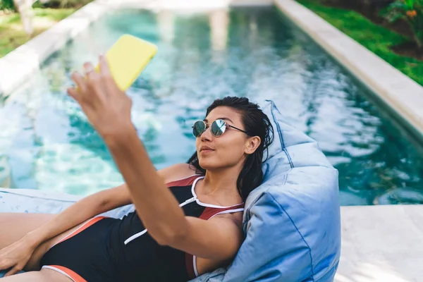 从上面看戴着太阳镜 身穿智能手机泳衣的女性 她们把自己放在豆袋躺椅上 并在暑假里放松一下 — 图库照片