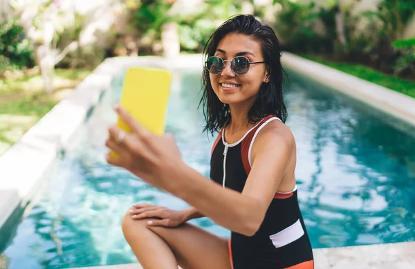 在阳光灿烂的日子里 身穿泳衣 头戴智能手机坐在游泳池边 一边自拍一边笑的迷人女性 — 图库照片