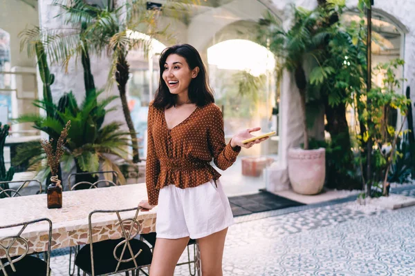 幸せな若いアジアの女性暗い髪でスタイリッシュな夏の衣装は椅子に手でテーブルの近くに立って スマートフォンを保持 — ストック写真