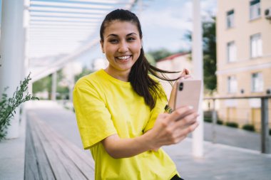 Neşeli etnik kadın, dişlek gülüşüyle ahşap bankta oturuyor ve akıllı telefondan selfie çekerken kameraya bakıyor.