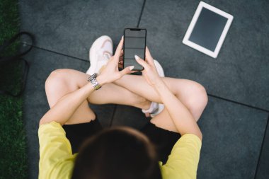 Spor kıyafetleri içinde koyu renk saçlı, bacak bacak üstüne atmış ve cep telefonuna mesaj atmış bir kadının en iyi görüntüsü.