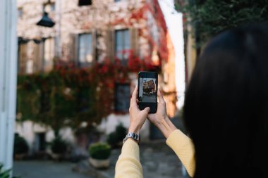 Kırpılmış kadın turist cep telefonu kamerasıyla güzel kasaba binalarına tıklıyor. Filtre uygulaması için 4G 'ye bağlanıyor. Görüntülü fotoğraf ile modern akıllı telefon teknolojisine odaklanıyor.