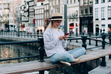 Günlük kıyafetleri ve şapkasıyla neşeli genç bir bayan Amsterdam 'da tatilde dinlenirken ahşap bankta oturuyor ve akıllı telefonla mesajlaşıyor.