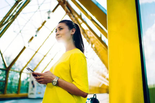電話を使用してインターネットを閲覧しながら離れて見て現代のガラス橋の黄色の鋼ビームの近くに明るい黄色のシャツを身に着けている若い女性の低角度 — ストック写真